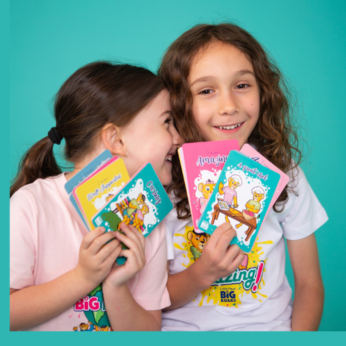 Little Paws Big Roars: Empowering Families Through Play Two children smiling and holding colourful Little Paws Big Roars cards with affirmations such as 'Amazing' and 'A Great Job'. The background is teal, creating a bright and cheerful atmosphere
