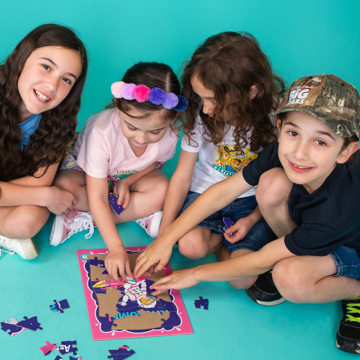 Little Paws Big Roars: Empowering Families Through Play Four children sitting on a teal background, collaborating on assembling a puzzle featuring a vibrant, illustrated image. The children are smiling and engaged, promoting teamwork and creativity