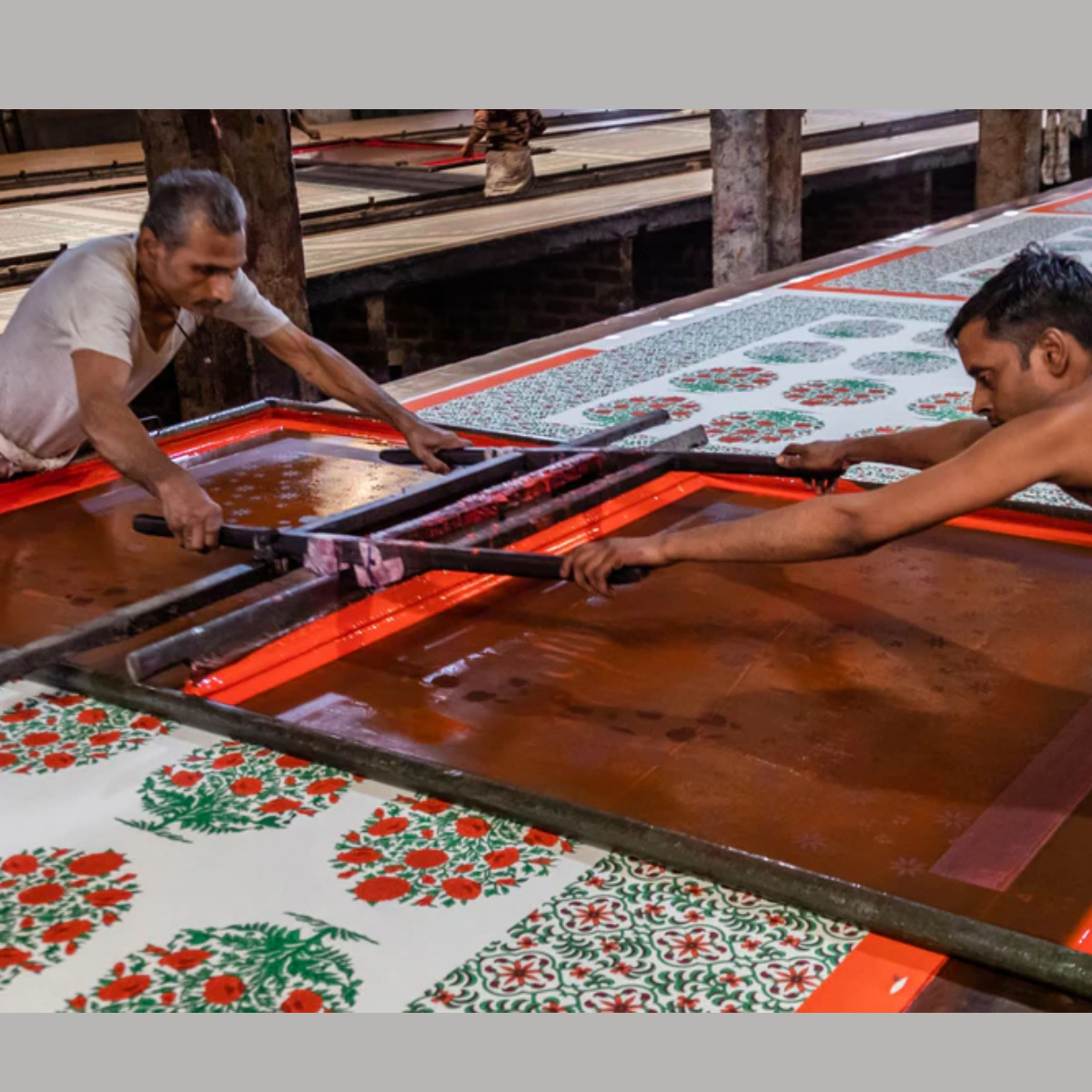 Greatest Good member feature hiHandmade Block printing process Block Printing Process: Two artisans working collaboratively on a traditional block printing process, using a large frame to print intricate floral and geometric patterns in vibrant colours on fabric. The setting appears to be a workshop with rows of printed textiles.