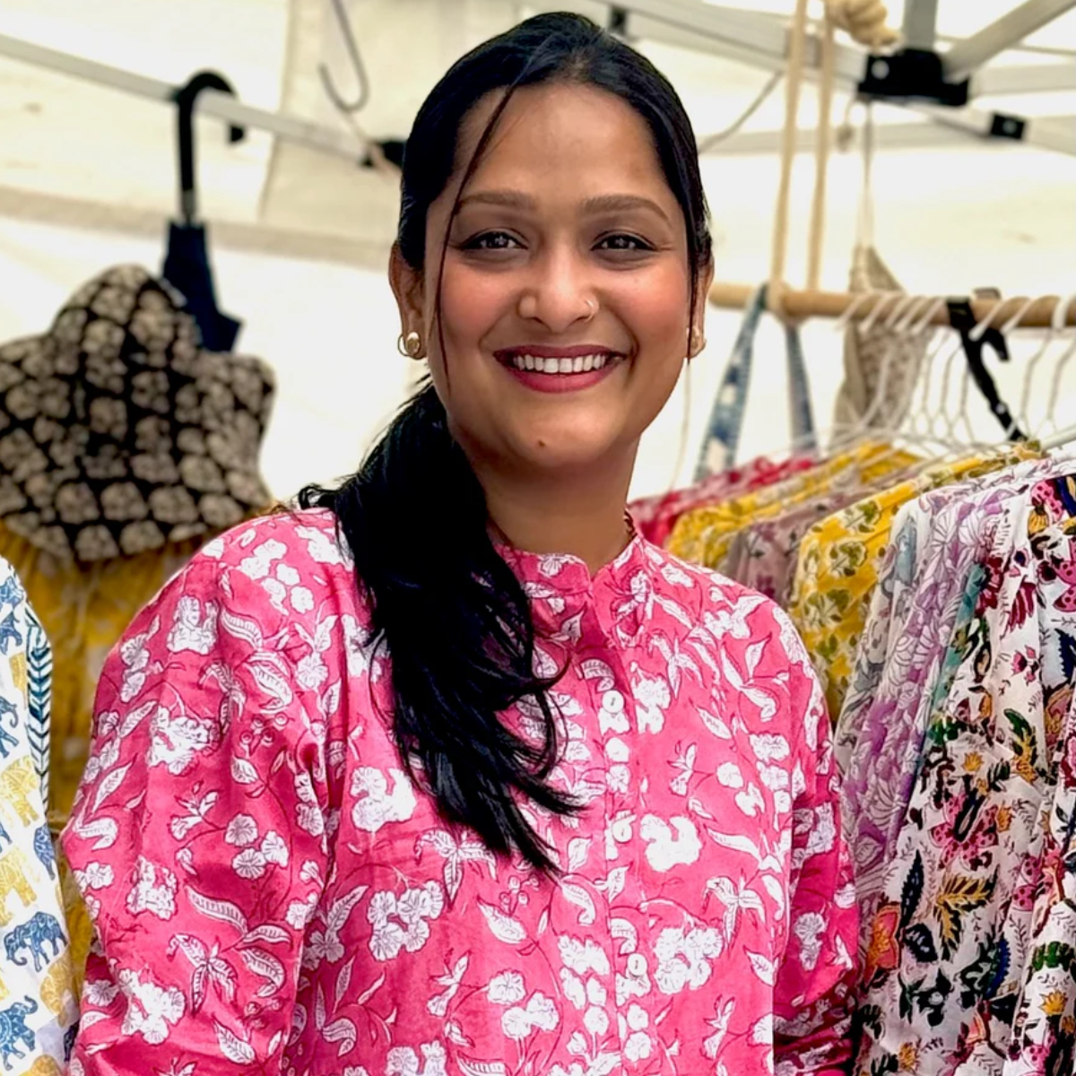Greatest Good Member Feature hihandmade artisan hiHandmade Artisan: A smiling woman wearing a bright pink floral top, standing amidst racks of colourful hand-printed garments. The photo captures a vibrant and cheerful market or boutique setting.