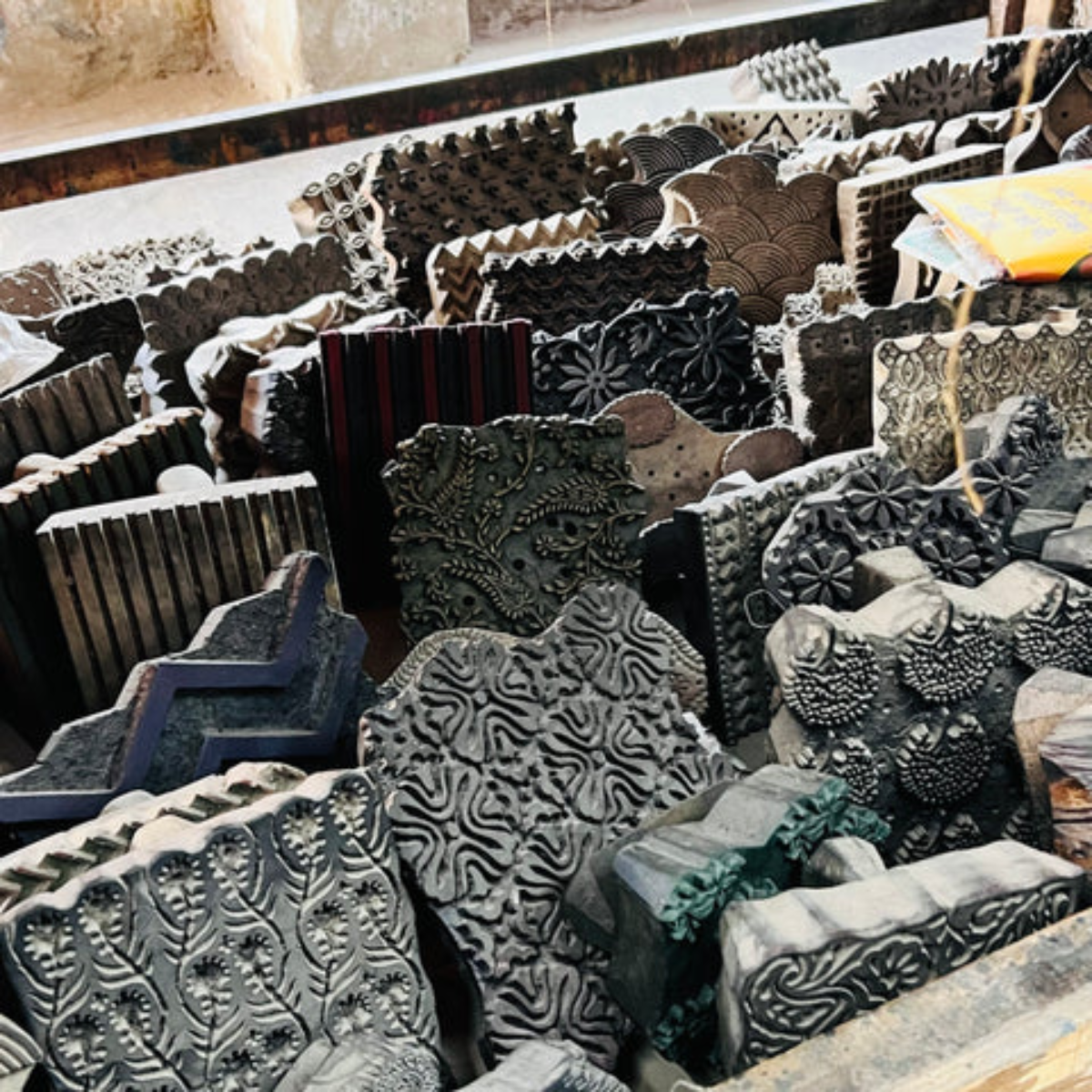 Greatest Good Member Feature hihandmade block printing stamps Block Printing Stamps: A collection of intricately carved wooden block stamps, used for traditional fabric printing. The blocks feature a variety of patterns, from floral to geometric, arranged in a workshop-like environment.