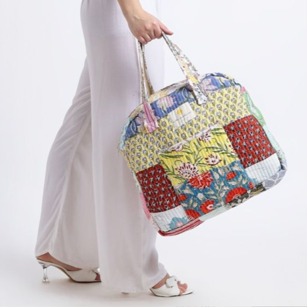 Greatest Good Member Feature hiHandmade quilted bag Quilted Bag: A patchwork quilted bag with vibrant floral and geometric patterns in shades of yellow, red, and blue, held by a person wearing white pants and heeled sandals. The bag reflects artisanal craftsmanship and a bohemian style.